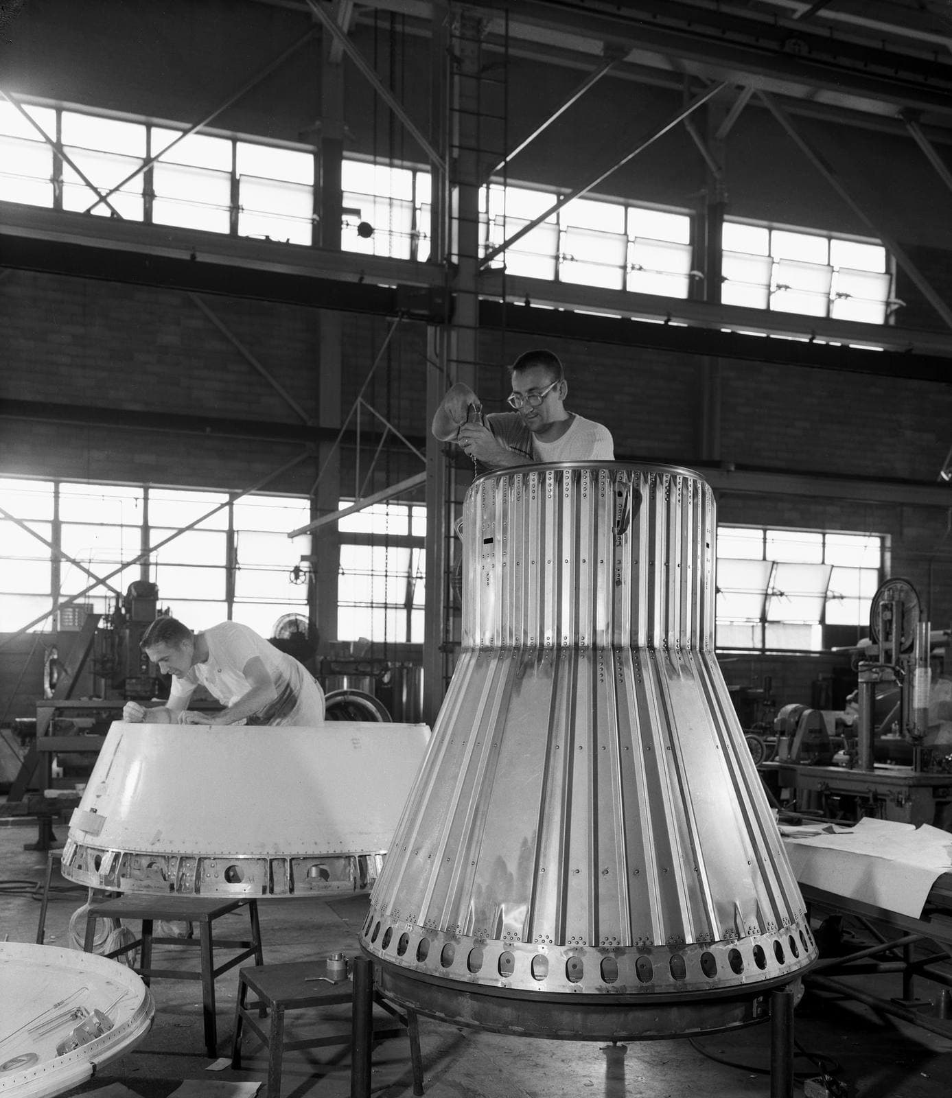 Two technicians in a high-bay hangar assembling cone-shaped Little Joe test capsules, one standing inside an open shell