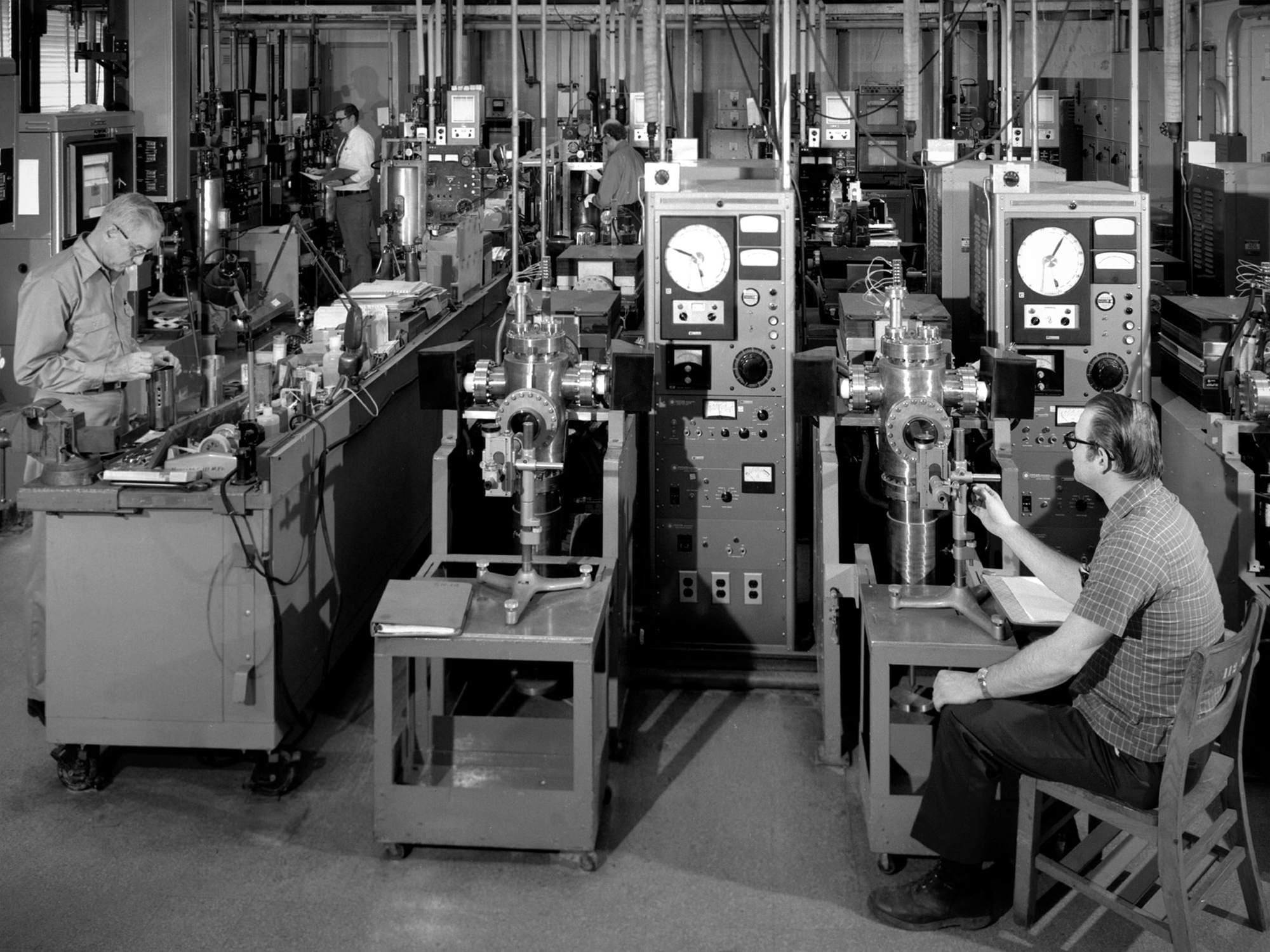 Row of technicians at instrument consoles with dial gauges and vacuum chambers in a 1970s materials test laboratory