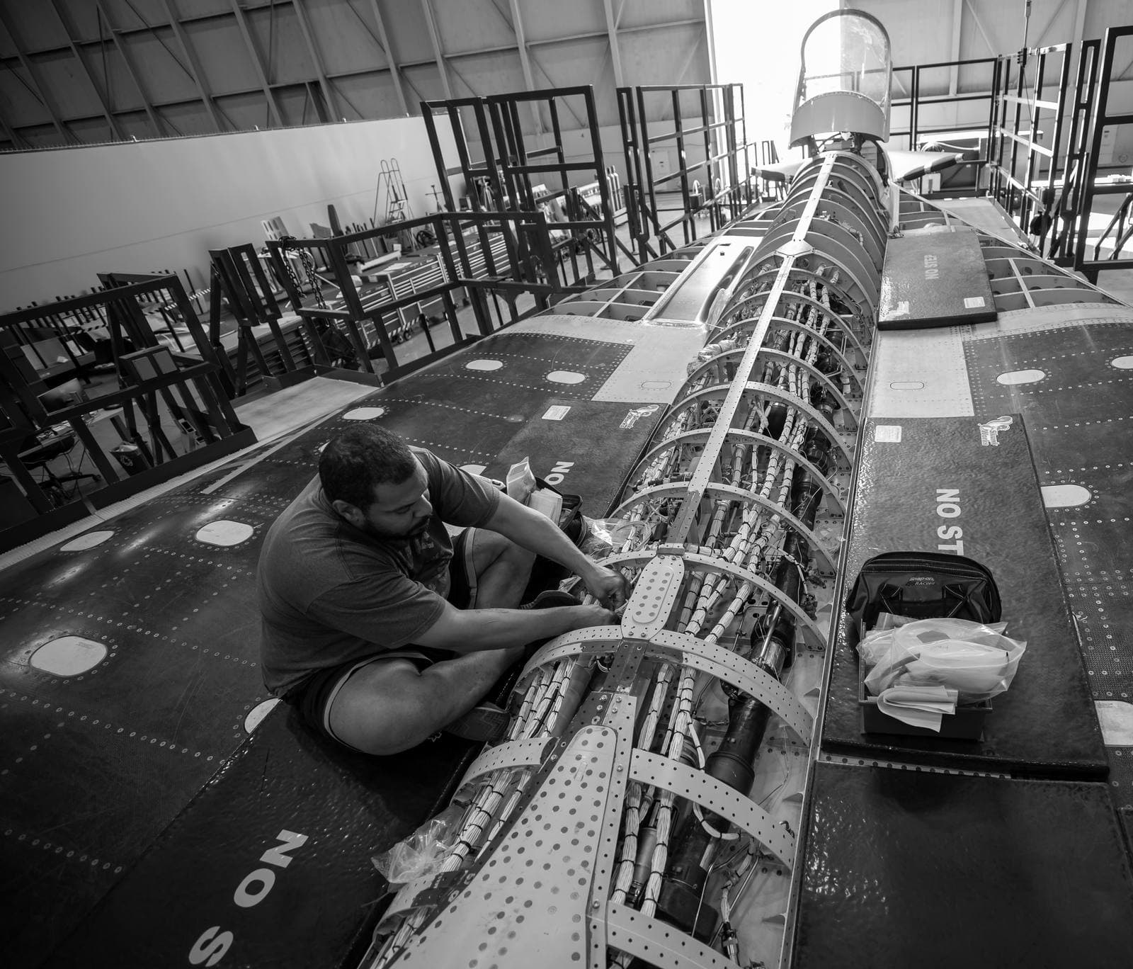 Technician sitting on an aircraft's upper fuselage, working on an open wiring bay running down the spine of the plane